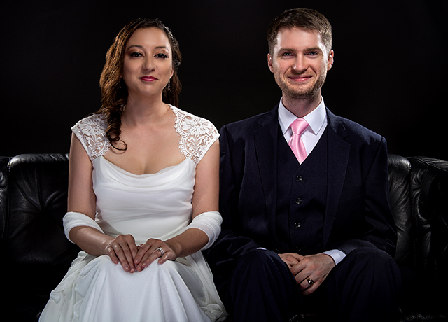Engaged couple wearing modern wedding suit and bridal dress in a classical retro fashion style.  The stylish groom and bride are sitting on a leather couch in a studio.